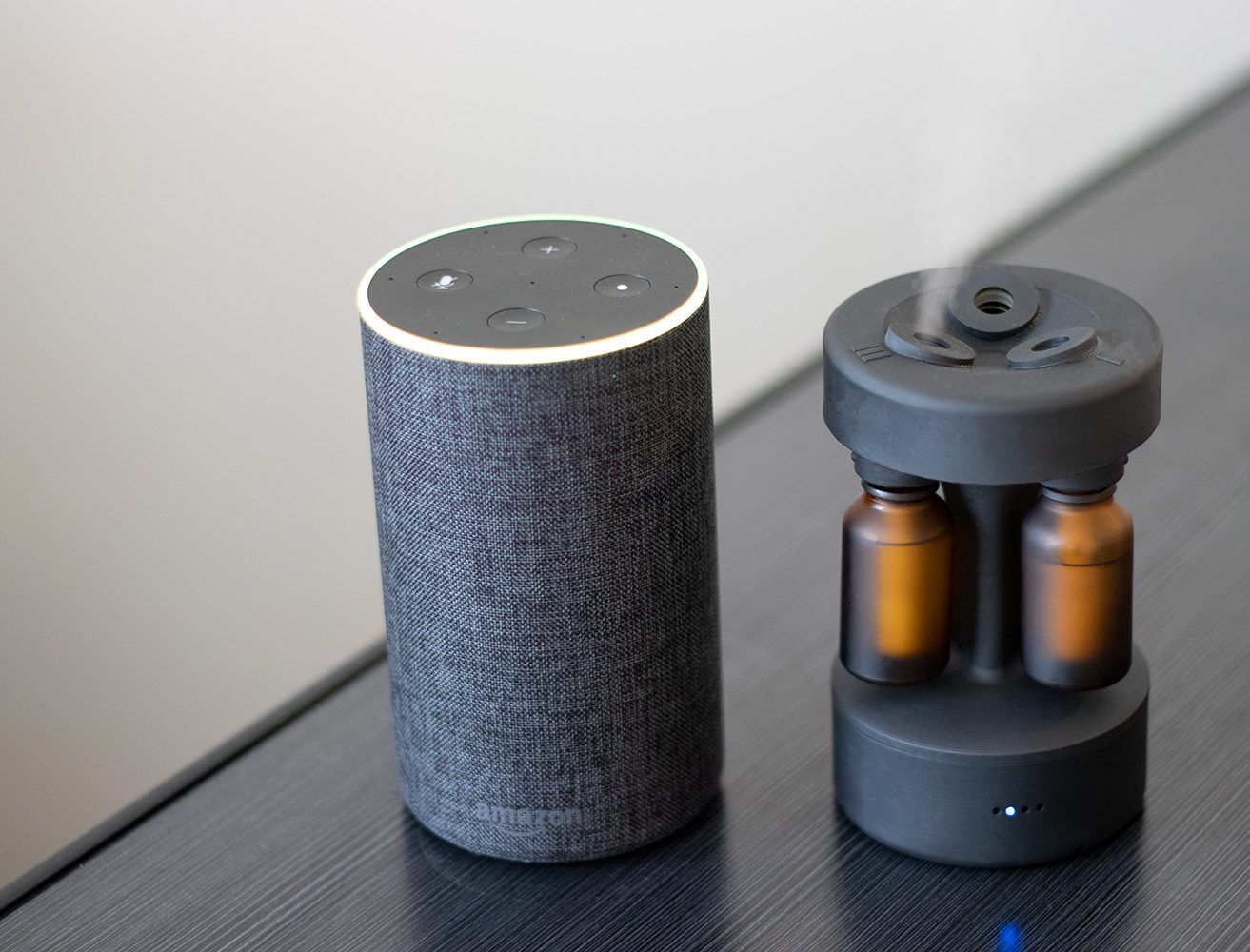 Curie diffuser sitting next to an Amazon Echo speaker on a dark table, the diffuser's blue LED visible, both devices roughly the same height.