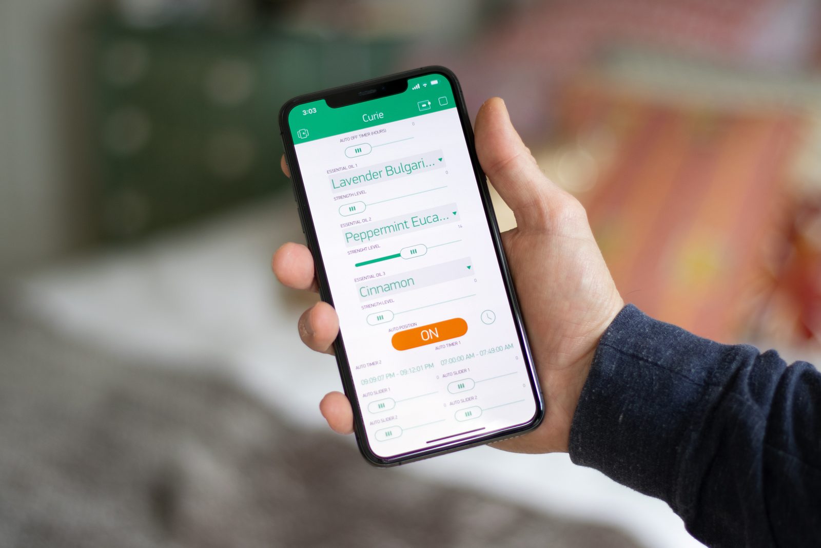 Hand holding an iPhone running the Curie iOS app, showing three essential oil slots — Lavender Bulgarian, Peppermint Eucalyptus, Cinnamon — each with its own strength slider and auto-timer schedule.