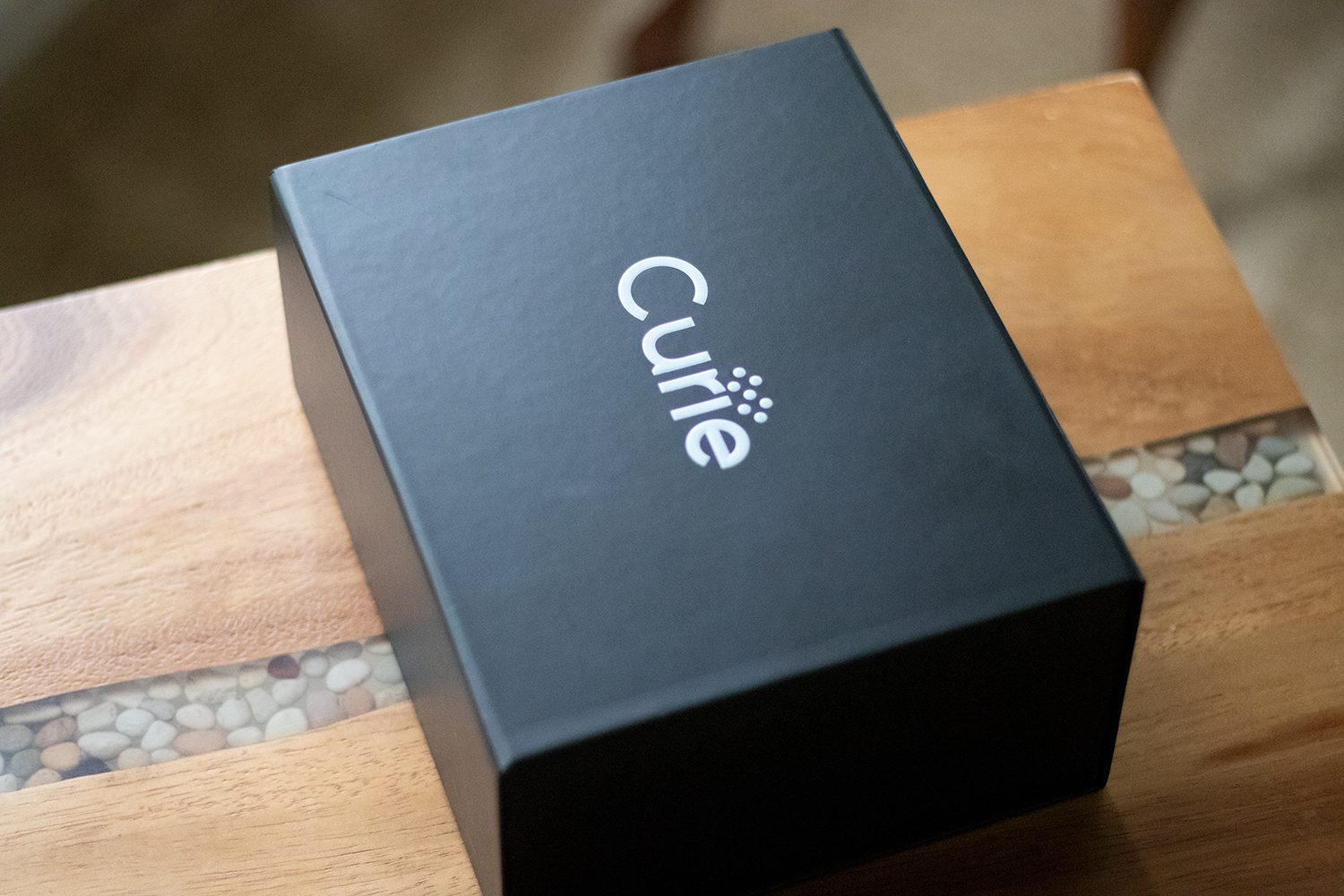 Curie retail packaging: a matte-black box with the Curie wordmark in white, resting on a wooden bench.