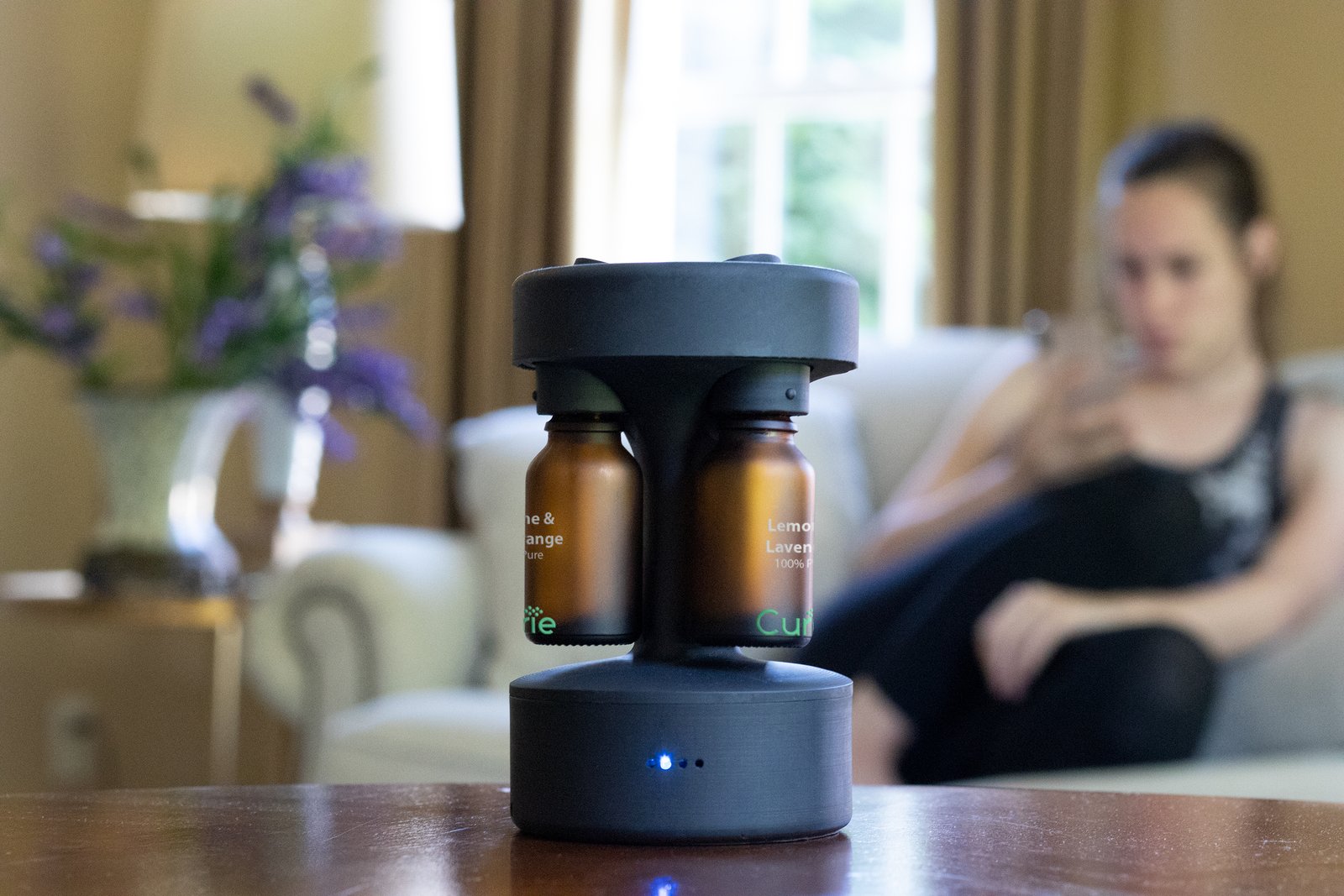 Curie diffuser on a living room table, amber bottles loaded, LED indicator lit, a person seated in the background.